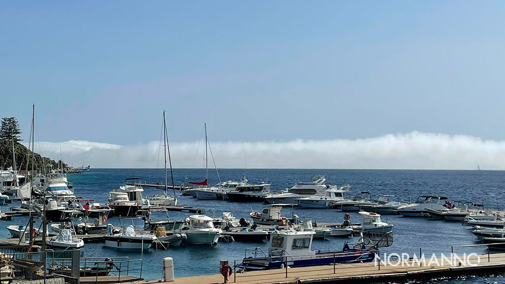 A low bank of fog crossing the Strait of Messina behind a marina, under a clear sky